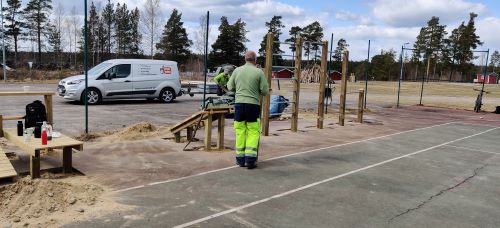 En person i arbetsklder gr frn kameran. I bakgrunden en skpbil med slp. En ryggsck, termosar och vattenflaskor str p situp-brdan.